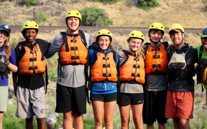 a group of students wearing helmets and life jackets stand and with their arms around each other and smile at the camera